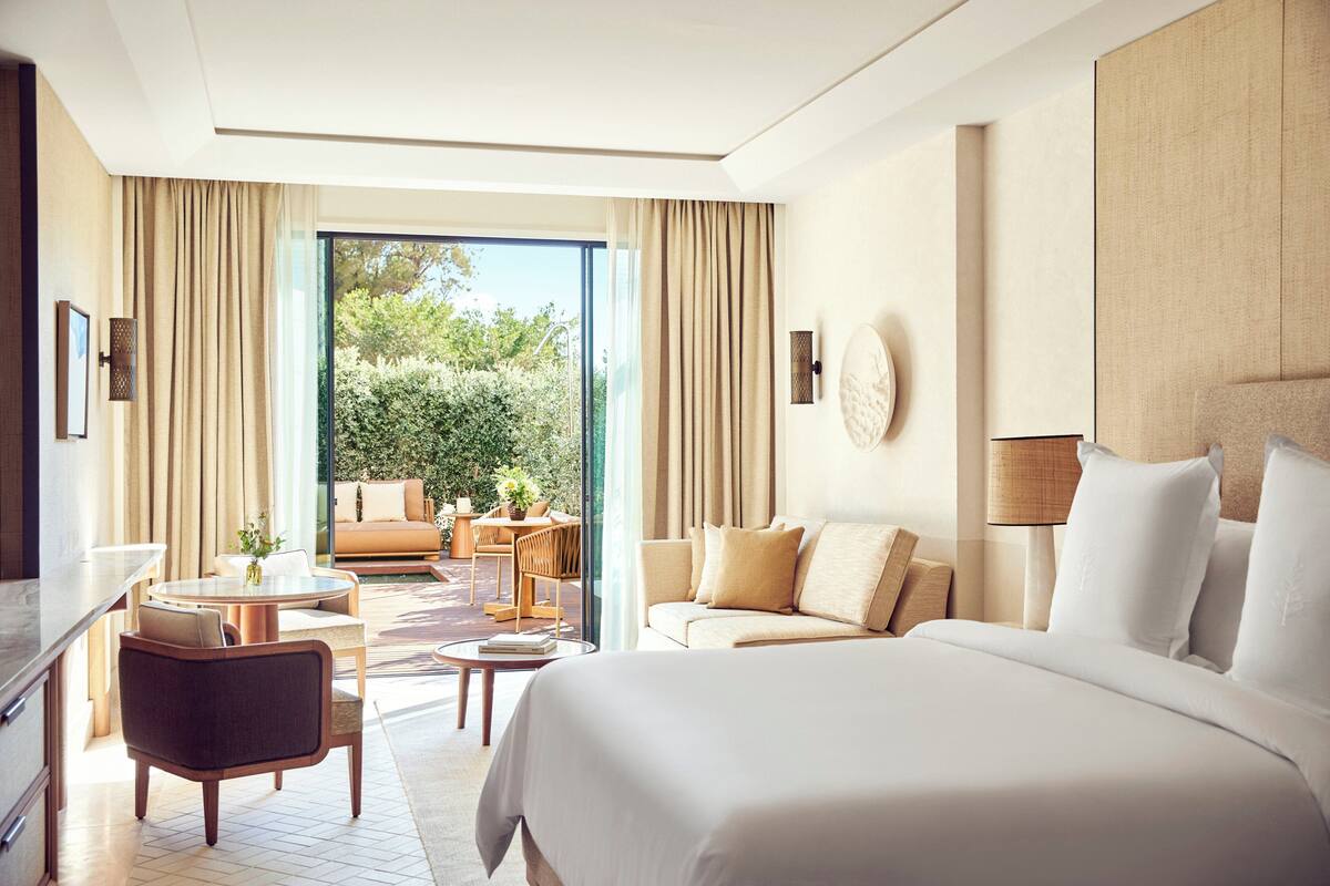 A hotel room with a large bed, a sofa, a dining area with a table and chairs, and a view of greenery outside.