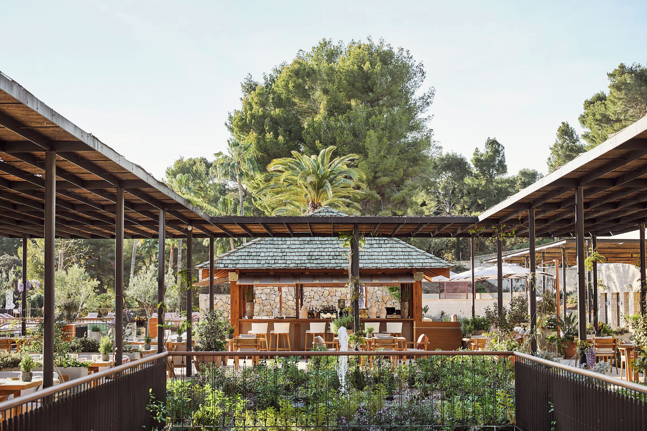 An outdoor seating area with wooden tables and chairs, a stone wall, and a pergola structure.