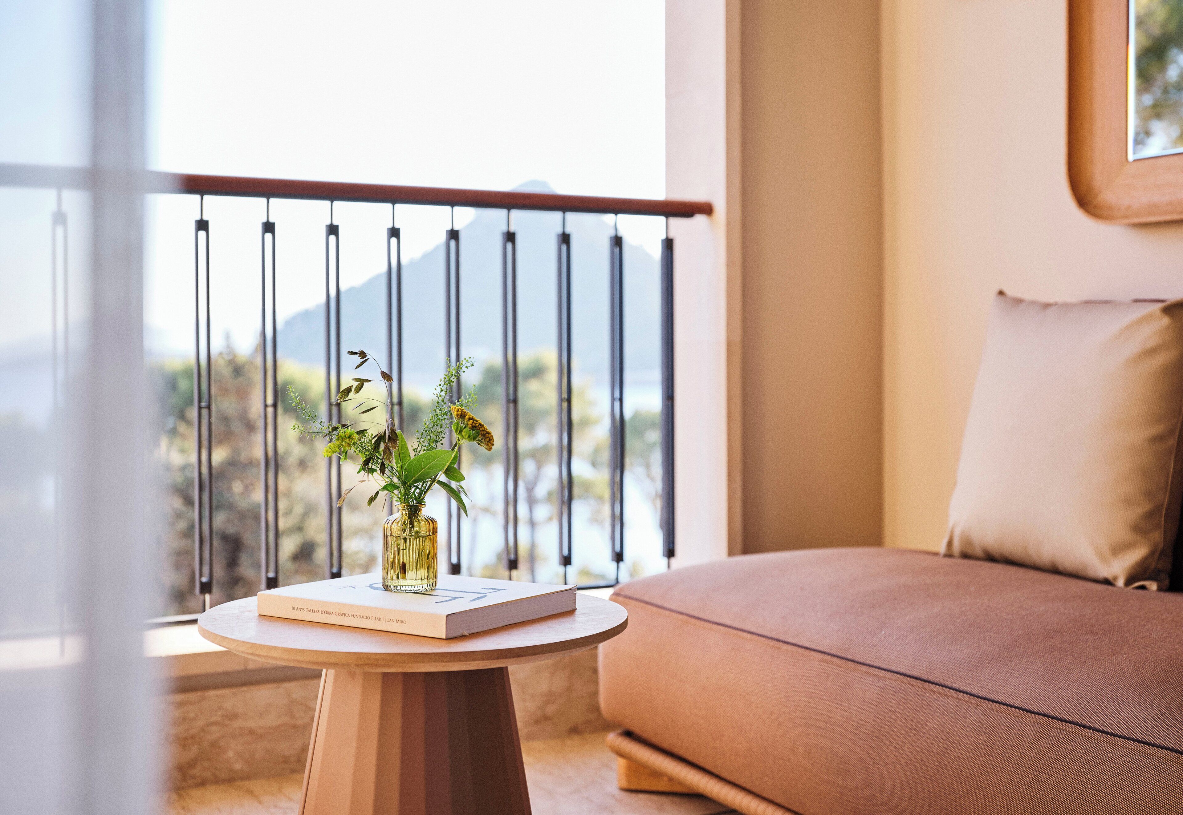 Una mesa redonda de madera con un libro y un jarrón de flores, un sofá marrón y una barandilla de balcón con vistas a la montaña.