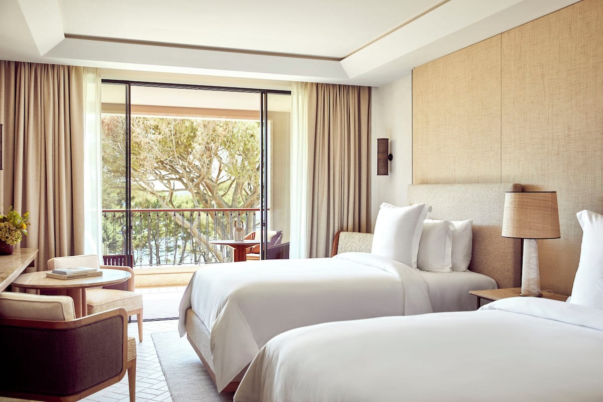 A hotel room with two beds, a desk, a chair, a lamp, and a view of trees through large windows.