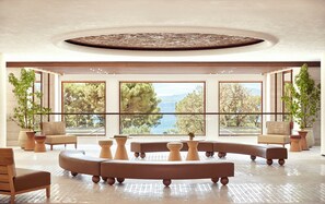 A spacious lobby with a large window offering a scenic view of trees and a body of water. There are several seating areas with round tables and chairs.