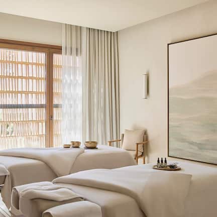 A spa room with two massage tables, a large window with wooden blinds, a painting on the wall, and a small table with bottles.