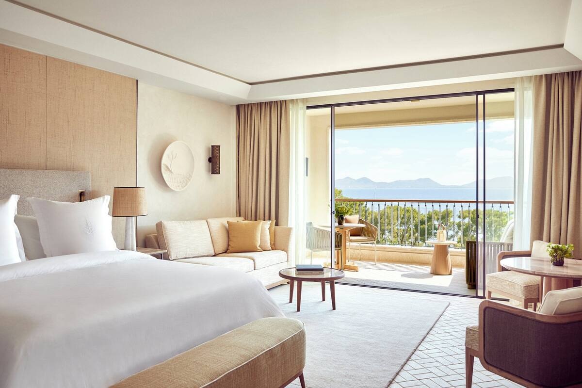 A hotel room with a large bed, a sofa, a balcony with a view of the mountains, and a dining area.