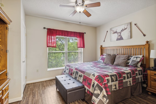 A bedroom with a bed, a nightstand, a window with blinds, a ceiling fan, and a wall-mounted picture.