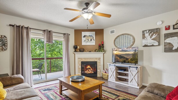 A living room with a fireplace, a ceiling fan, and a large window with curtains. - Stay on the River, Steps from Dolly Parton’s Stampede – Charming 2BR Retreat (Pigeon Forge)