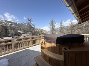 Une terrasse en bois avec un jacuzzi et un banc en bois, offrant une vue sur un paysage montagneux enneigé.