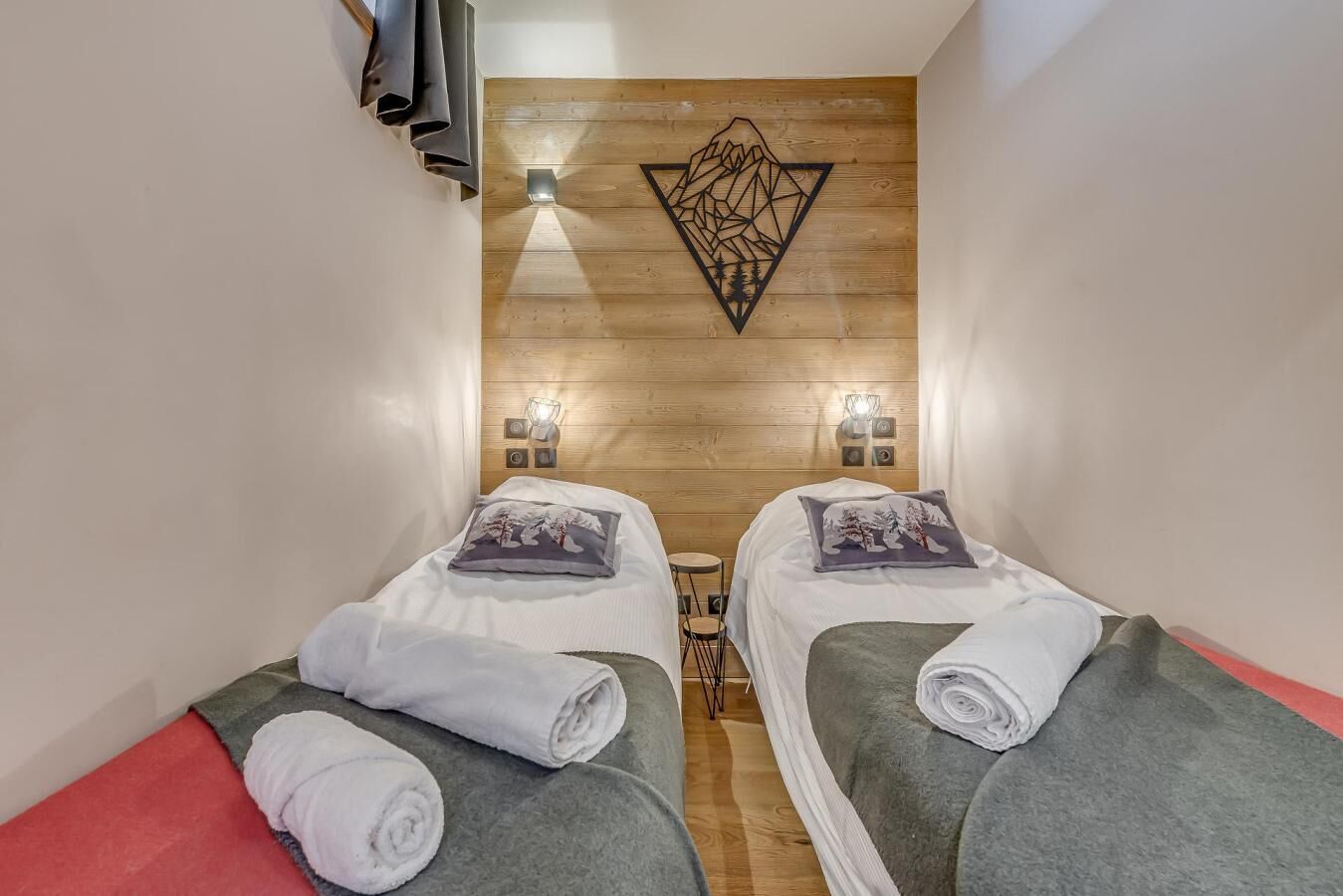 Una habitación con dos camas, cada una con una almohada y una toalla enrollada. Hay luces fijadas en la pared y un panel de madera con un diseño geométrico.