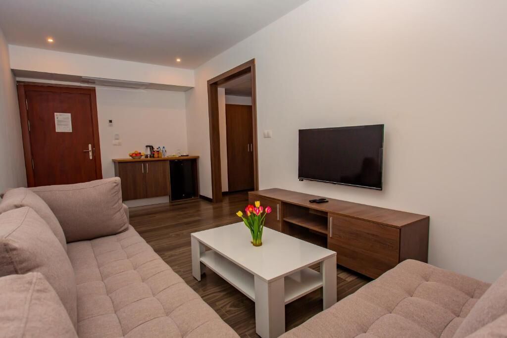 Una sala moderna con un sofá color crema, una mesa de centro blanca con un jarrón de flores, un televisor de pantalla plana montado en la pared y un mueble de madera.