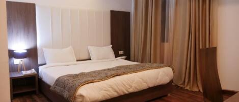 A hotel room with a large bed, wooden flooring, a nightstand with a lamp, and a view of the outside through curtains.