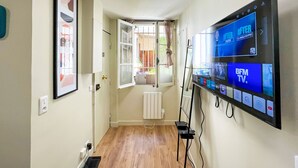 A hallway with a large flat-screen TV mounted on the wall displaying various channels. There is a window with a curtain, a radiator, and a door leading to another room. - Urban Flat 162 - Charming Studio in Montorgueil (París)