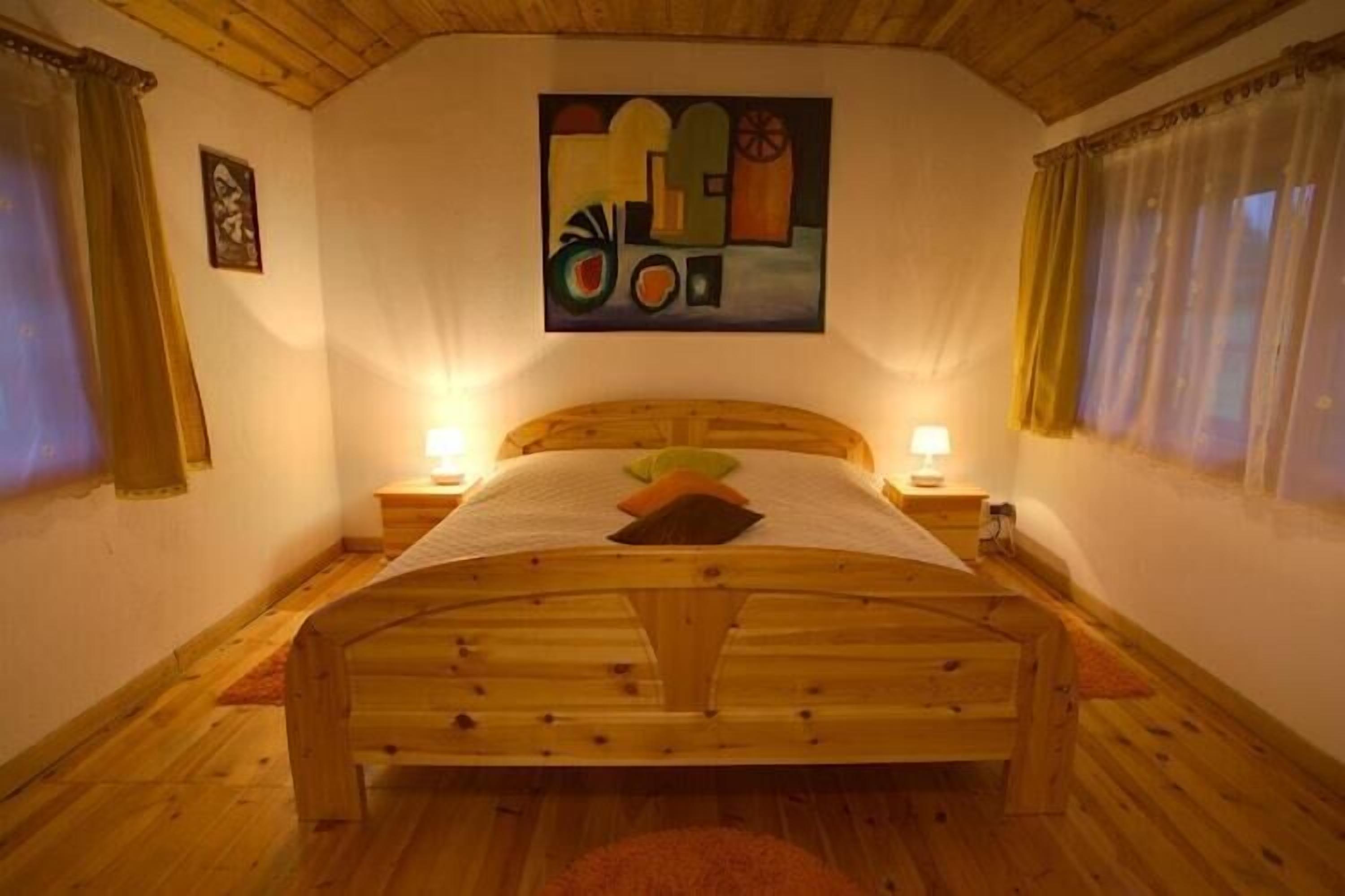 Uma cama de madeira com cabeceira, duas mesas de cabeceira com abajures, um quadro na parede e uma janela com cortinas.
