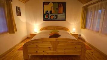 Uma cama de madeira com cabeceira, duas mesas de cabeceira com abajures, um quadro na parede e uma janela com cortinas.