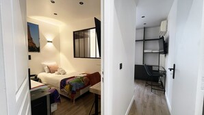 Quarto de hotel pequeno com cama, mesa de cabeceira e escrivaninha com cadeira. Há uma janela com vista para um edifício, e uma prateleira fixa na parede com uma cadeira.