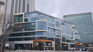 Um edifício moderno com grandes janelas de vidro, uma entrada destacada e uma área de estacionamento em frente.