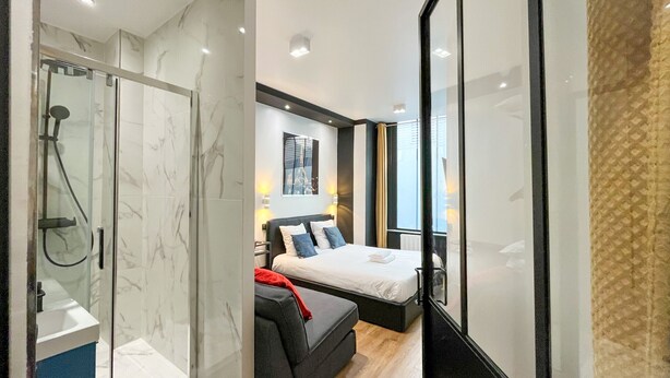 A modern hotel room with a glass shower enclosure, a bed with a black headboard, and a large window offering a view. - Urban Flat 173 - Atelier Gaite Montparnasse Apartment (París)
