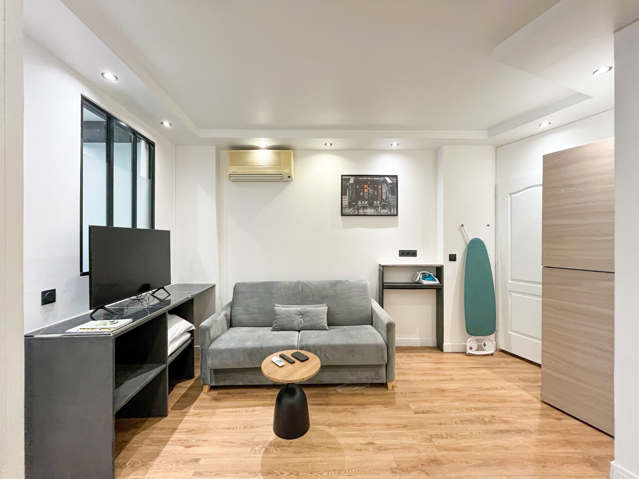 A modern living room with a grey sofa, a small round coffee table, a TV on a stand, and a wall-mounted air conditioner.