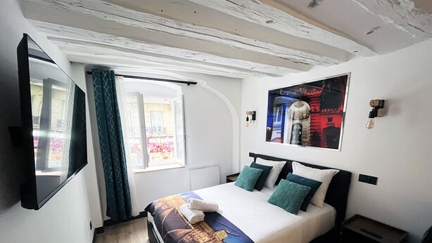 Un dormitorio con cama, ventana, televisor montado en la pared y un cuadro enmarcado.