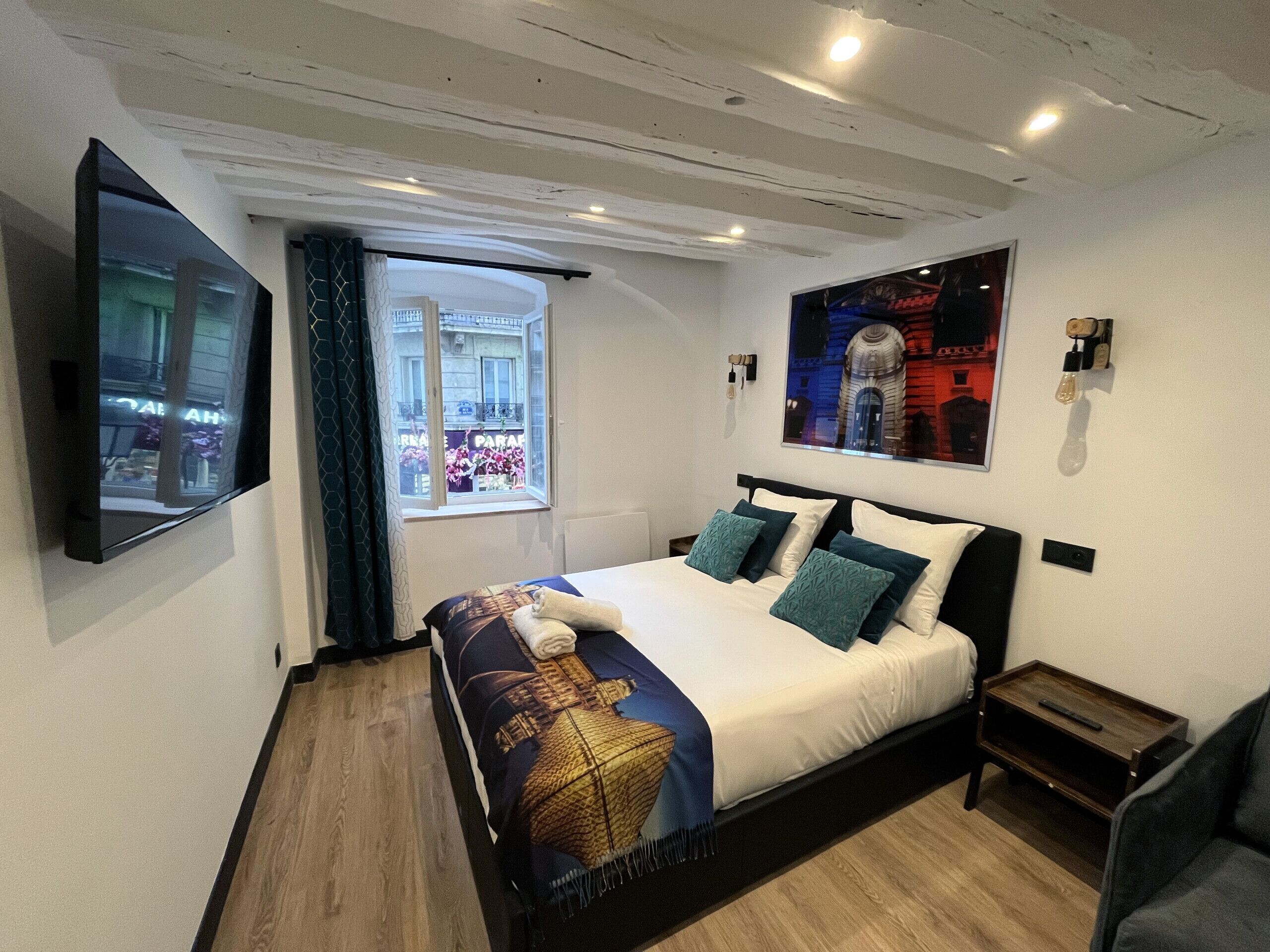 A modern hotel room with a large bed, a TV mounted on the wall, a window with a view of the city, and a framed picture on the wall.