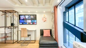 Ein modernes Hotelzimmer mit Bett, Schreibtisch, Fernseher, der verschiedene Sender zeigt, und einem großen Fenster mit Blick auf die Stadt.