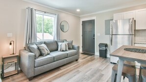 Un apartamento moderno con un sofá gris, una mesa de comedor con sillas, una cocina con un refrigerador de acero inoxidable y un amplio ventanal con cortinas.