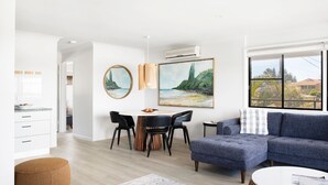 Un salon moderne avec un canapé bleu, un coin repas doté d’une table ronde et de chaises, et une cuisine avec des meubles blancs.