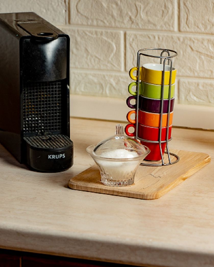 A coffee maker, a stack of colorful mugs, and a glass container on a wooden board.
