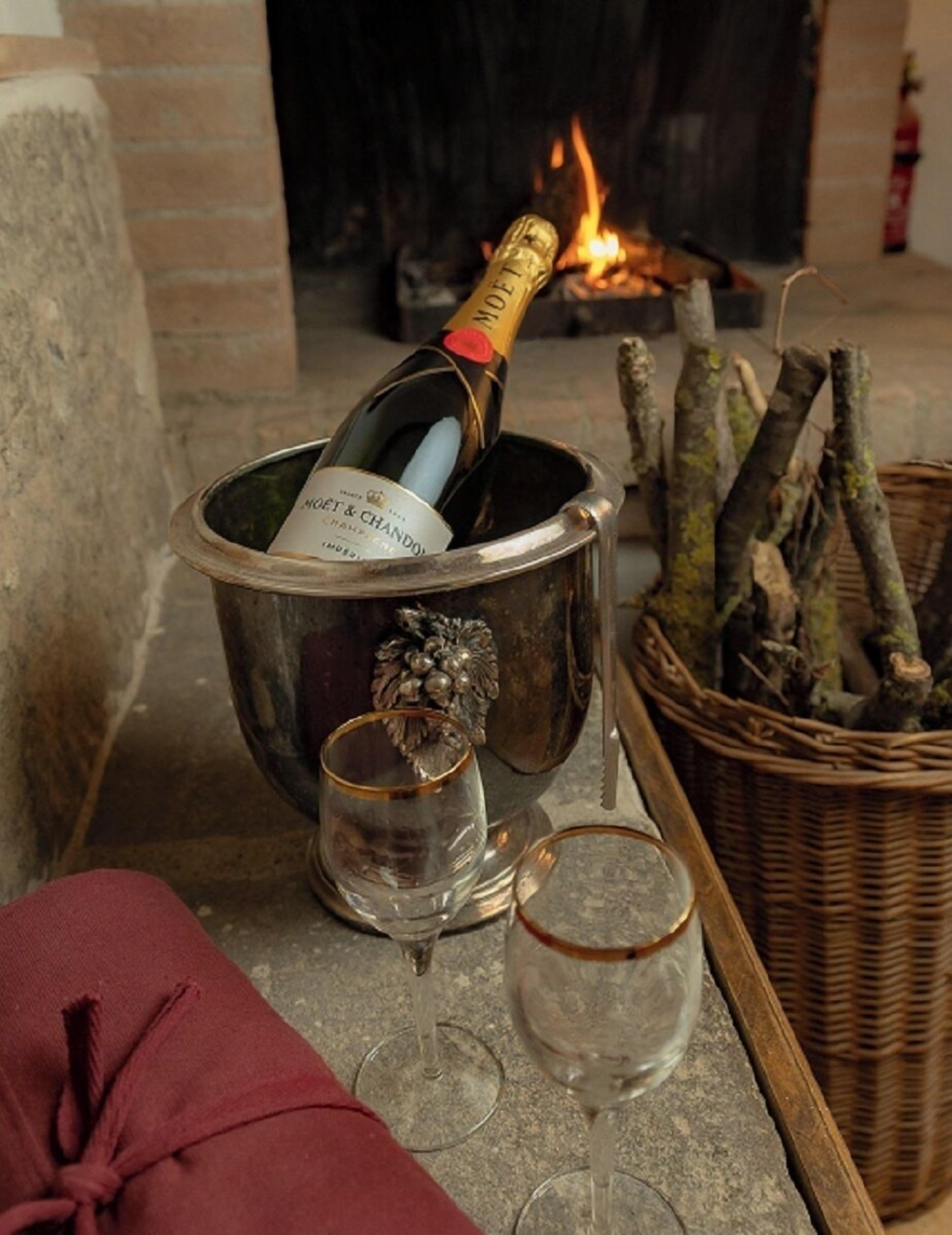 Una chimenea con una botella de champán en un cubo de metal, dos copas y una cesta de mimbre con leña.