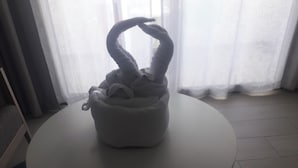 Una mesa blanca y redonda con un objeto suave de color gris, con forma de animal retorcido encima, sobre un fondo de cortinas translúcidas y un suelo de baldosas.