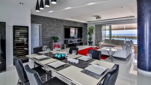 A modern dining area with a long table set for six, a flat-screen TV mounted on the wall, and a view of the sea through large windows. - Lovely 3-bed Apartment in Maho (Lowlands)