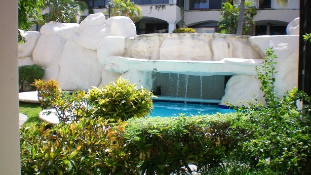 A pool area with a waterfall feature, surrounded by greenery and a building in the background. - Casa Clarita top Location for Your Cancun Experience (Cancún)