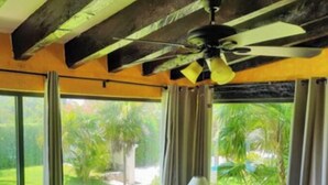 A room with two tufted armchairs, a wooden nightstand, a lamp, and a ceiling fan. Large windows offer a view of a green landscape. - Casa Clarita top Location for Your Cancun Experience (Cancún)
