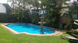 Una piscina con fuente, rodeada de césped cuidado y plantas tropicales.