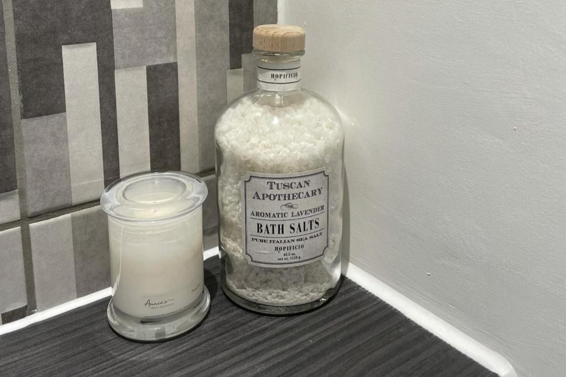 Une bouteille de sels de bain aromatiques à la lavande de la marque Tuscan Apothecary et une bougie posées sur une étagère.