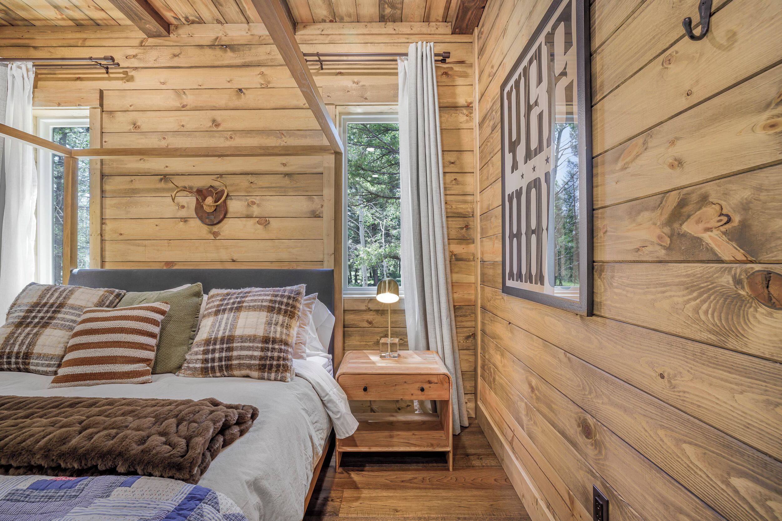 Une chambre à coucher dans une cabane en bois, avec un lit, des oreillers, une table de chevet, une lampe et une fenêtre avec des rideaux.