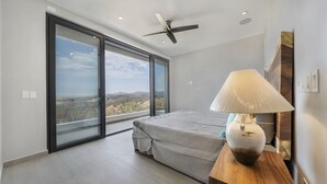 A bedroom with a bed, a ceiling fan, a nightstand with a lamp, and a large window offering a view of the landscape. - Casa Lukaz Penthouse private rooftop (San José del Cabo)