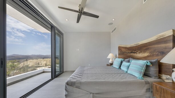 A modern bedroom with a large bed, a ceiling fan, and a view of the desert landscape through large windows. - Casa Lukaz Penthouse private rooftop (San José del Cabo)