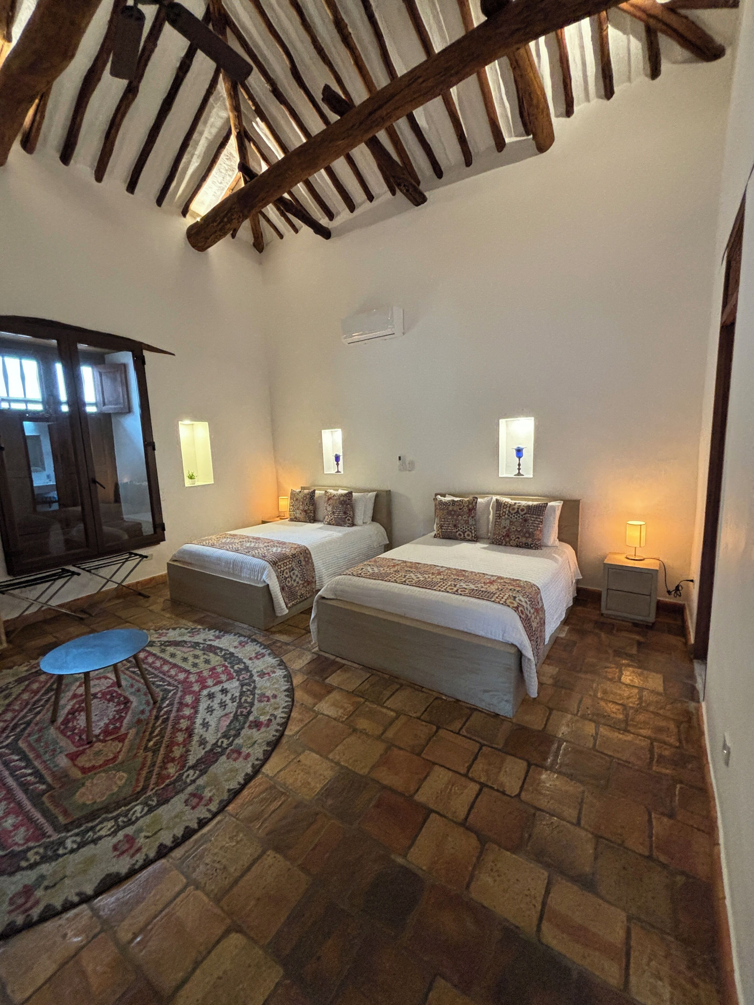 A cozy bedroom with two beds, a small table, a patterned rug, and a wooden ceiling.