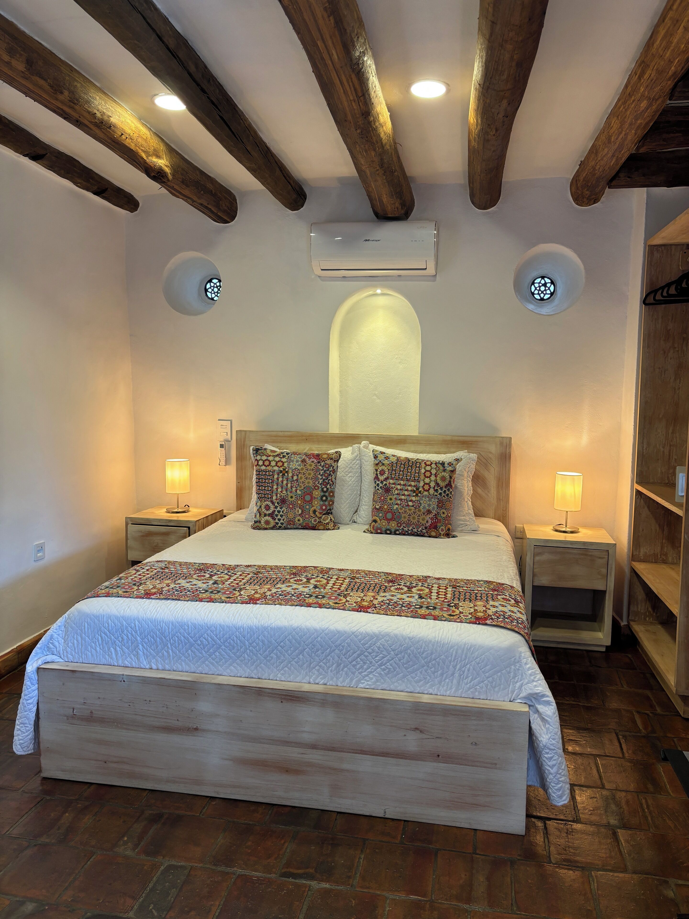 Een slaapkamer met een houten hoofdbord, een geborduurde spreideken, gestreepte kussens, twee bedlampjes en een luchtverkoeler aan de muur.