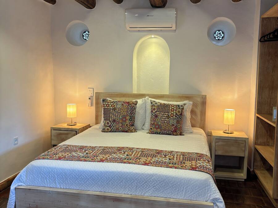 A bedroom with a wooden headboard, a quilted bedspread, patterned pillows, two bedside lamps, and a wall-mounted air conditioner.