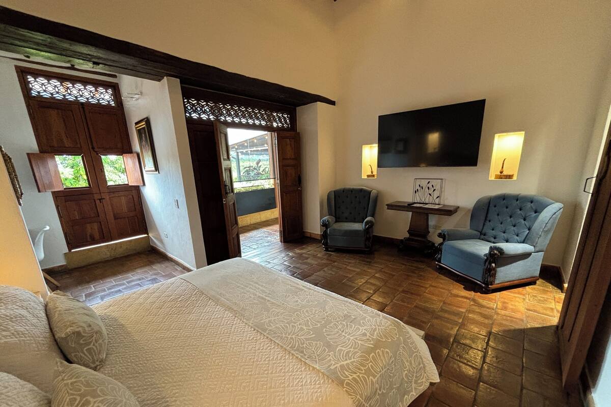Une chambre avec un grand lit, deux fauteuils, une petite table et un téléviseur fixé au mur.