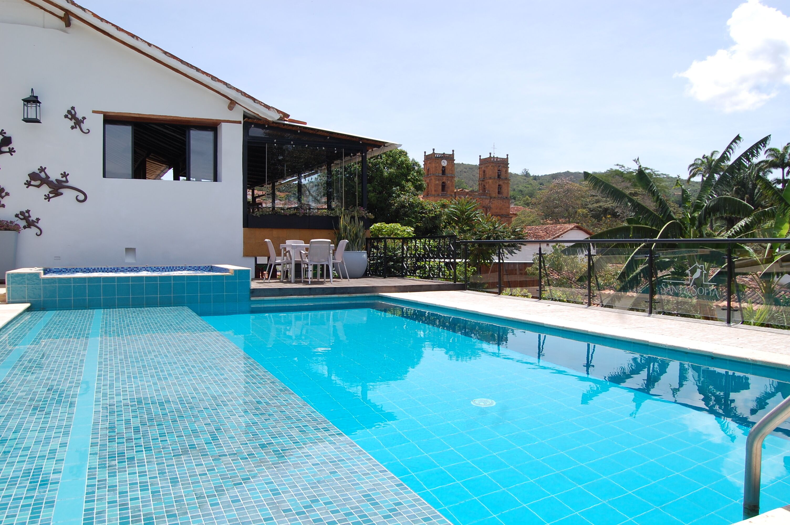 Una piscina con suelo de baldosas, un área de estar techada con sillas y una vista de una torre de iglesia al fondo.
