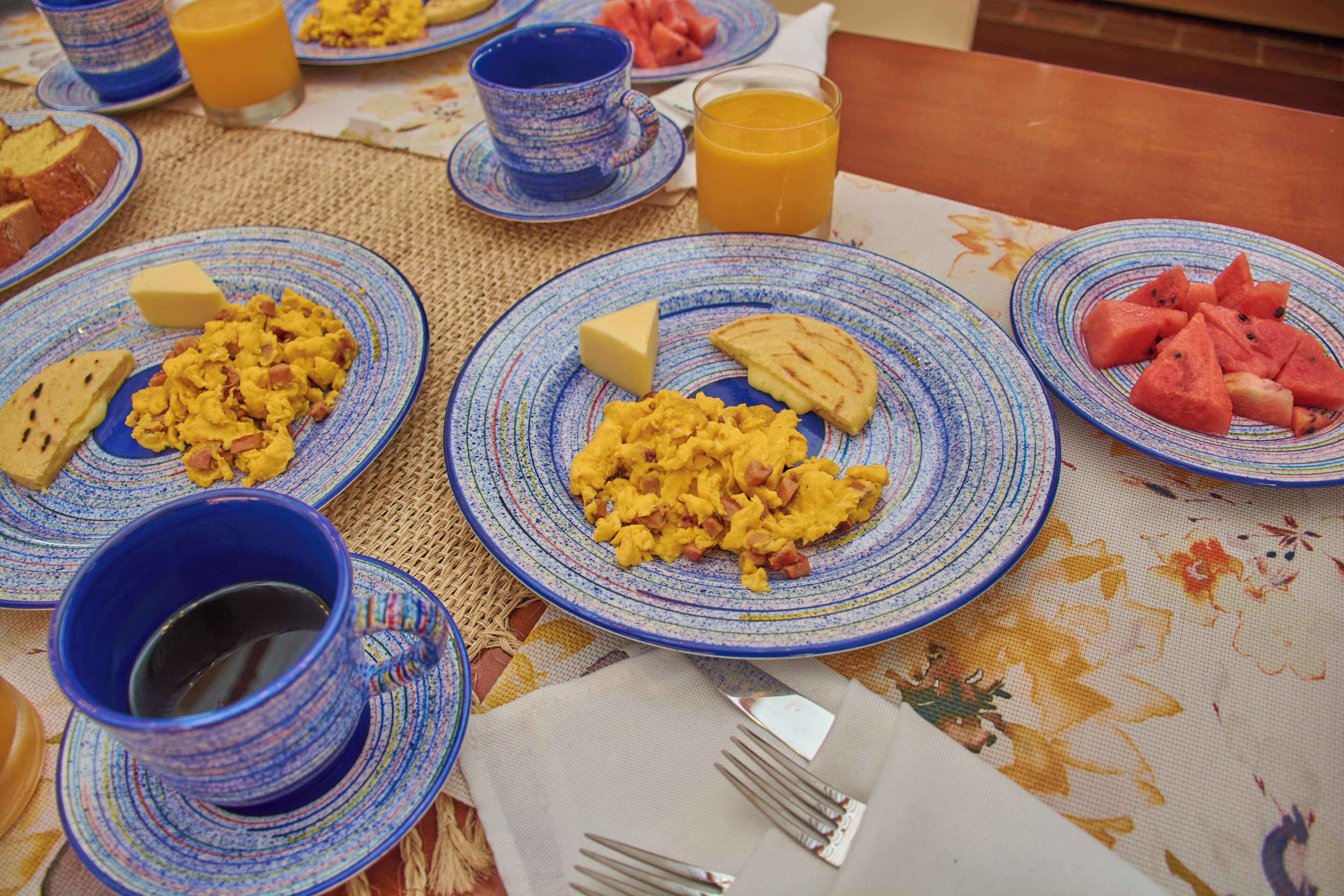 Una mesa de desayuno con platos de huevos revueltos, tostadas y sandía, además de tazas azules y un vaso de jugo de naranja.