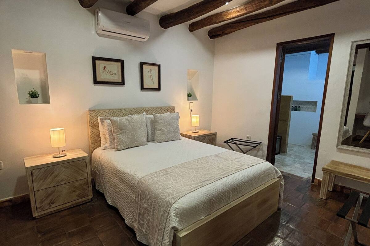 Une chambre avec un grand lit, des poutres en bois au plafond, une petite table de chevet avec une lampe et un miroir au mur.