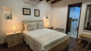 A bedroom with a large bed, wooden beams on the ceiling, a small bedside table with a lamp, and a mirror on the wall.