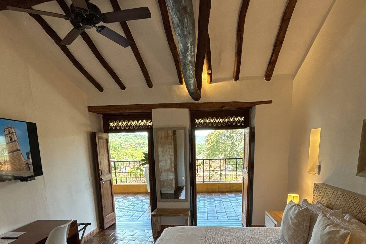 Une chambre avec un grand lit, un bureau et une chaise. On y trouve un ventilateur de plafond et une vue sur l’extérieur par une porte.