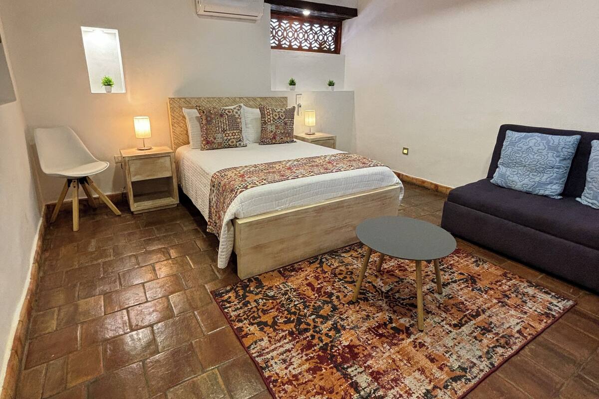 Une chambre à coucher comprenant un lit, des tables de chevet, une petite table, un canapé, un tapis à motifs et des poutres apparentes en bois au plafond.