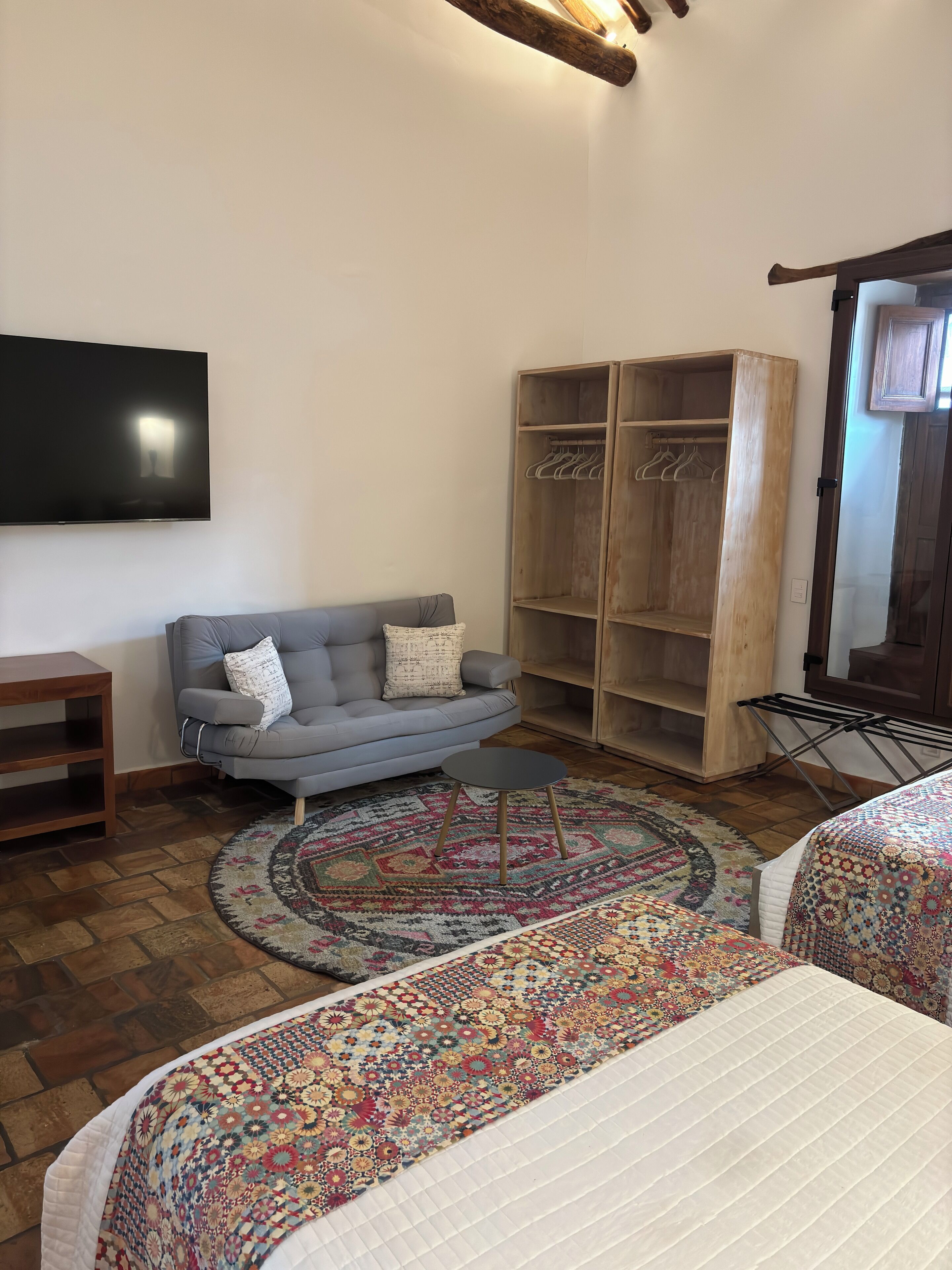 A cozy living room with a grey sofa, a wooden wardrobe, a flat-screen TV, and a patterned rug.