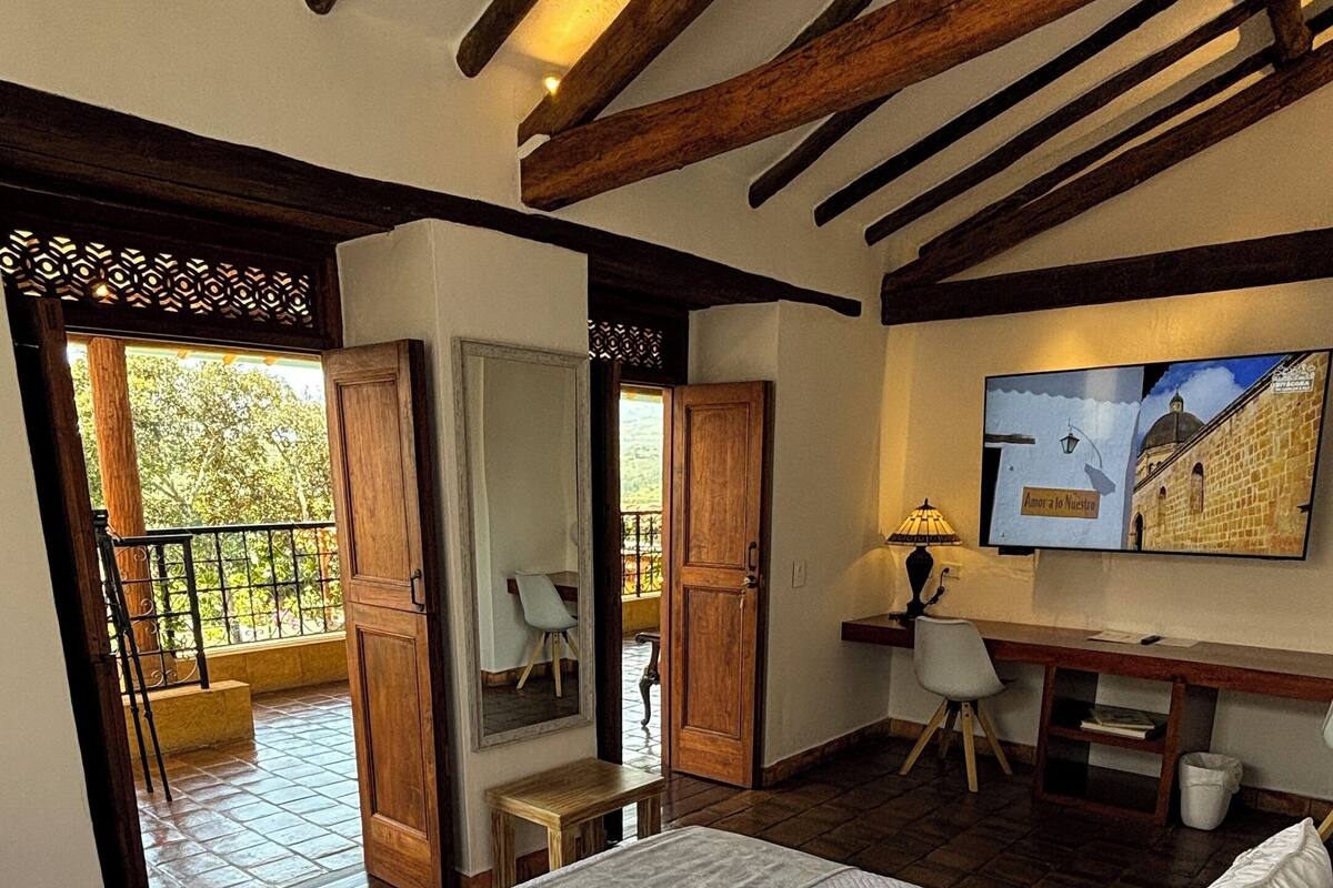 Une chambre avec un grand lit, des poutres apparentes, un bureau et une vue sur l’extérieur.