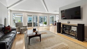 A spacious living room with a leather sofa, a patterned armchair, a coffee table, and a fireplace. A large TV is mounted on the wall, and there is a sliding glass door leading to an outdoor area.