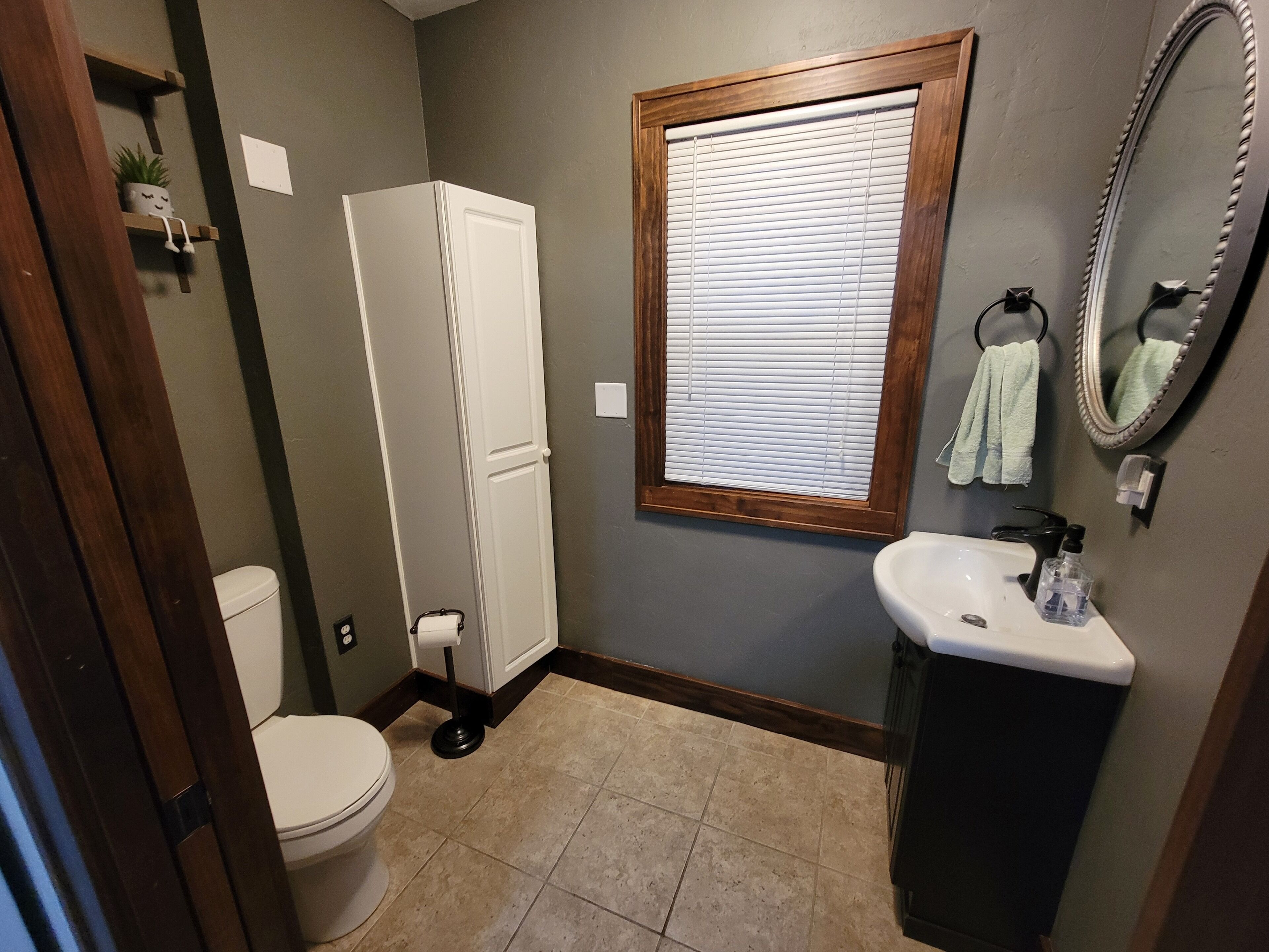 A bathroom with a toilet, a white cabinet, a sink, a mirror, and a window with blinds.